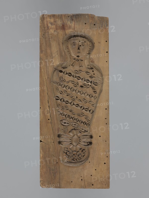 Double-sided gingerbread form. Obverse: female figure in a hat and a long patterned dress. Reverse: baby in swaddling clothes, richly decorated with geometric and plant ornament. Rectangular form; decorative art/folk customs motif. 18th–19th century. Province of Silesia 1742–1918. Height 23.5 cm; width 9.5 cm; depth 2.6 cm., A vertical rectangular wooden plank in brown tones with a carved recessed figure occupying the center, the carving depicting a stylized upright humanlike form with a rounded head, simple facial features, and a hood or hair outline, the torso area filled with repeating leaflike and geometric carved patterns and a bowlike carved element near the lower portion above a floral motif, the wood showing darker and lighter brown grain variations, small round holes scattered across the surface, a few surface cracks and worn edges, and a neutral gray background.