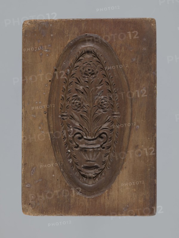 Double-sided gingerbread mould. Obverse: vase with flowers in an oval frame surrounded by a wreath of leaves. Reverse: rectangular recess with a plant ornament, rose motif and initials E and G. Rectangular shape; mould. 18th–19th century, Province of Silesia 1742–1918. Height 16.5 cm, width 11.5 cm, depth 2.7 cm., A rectangular wooden panel with a carved oval medallion at its center containing a symmetrical floral motif; the carving shows stylized flowers and leaves rising from a decorative vase or urn, with flowing curved elements and a textured carved border around the oval. The wood shows a range of brown tones with lighter and darker areas, small pale spots, and worn edges, and the background behind the panel is a muted gray.