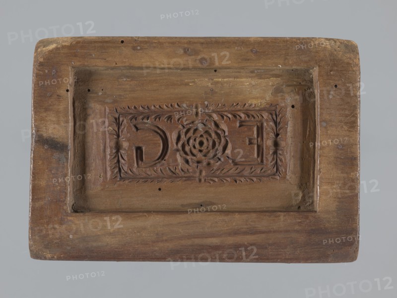 Double-sided gingerbread mould. Oblong rectangular mould; obverse: oval frame containing a decorative vase with flowers surrounded by a wreath of leaves; reverse: rectangular recess with a rectangular frame formed by a plant ornament featuring a rose motif and the initials E and G. Applied art, folk motifs, floral decorations; used for gingerbreads and wreaths. 18th–19th century, Province of Silesia 1742–1918. Mould. Height 16.5 cm, width 11.5 cm, depth 2.7 cm., A rectangular wooden panel with a recessed central area containing a carved rectangular cartouche; the cartouche has a carved floral rosette at its center and leaflike border carving surrounding it, with a few small round holes and surface blemishes across the wood; the panel shows a range of brown tones in the wood grain, with areas of lighter tan and some darker brown patches, set against a pale gray background.