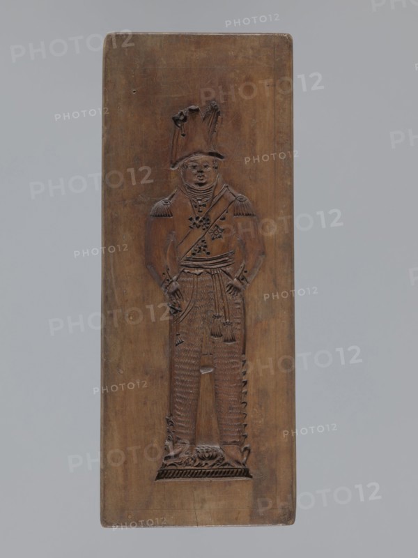 Double-sided gingerbread mould. Side A: figure of a soldier; Side B: various floral and geometric ornaments., woodcarver or gingerbread maker. Folk-style decorations; woodcarving; applied and decorative art; gingerbread moulds; male figures; military uniforms; folk motifs. Double-sided rectangular mould: one side shows a male figure in a Napoleonic-era military uniform, wearing a high headgear with a feather, numerous chest decorations, and a plant ornament at the bottom; the other side shows a rich floral and plant ornament enclosed in a tripartite frame with decorative spoked wheels in the four corners. On one edge an engraved date 1810 appears, with the letter S between the digits against the background of an anchor the maker's mark. Date: 1810. Place: Province of Silesia 1742–1918. Object type: form. Dimensions: height 31.7 cm; width 12.5 cm; depth 2.4 cm., A rectangular wooden panel with a carved standing figure in relief occupies the center; the panel is brown with variations in ...