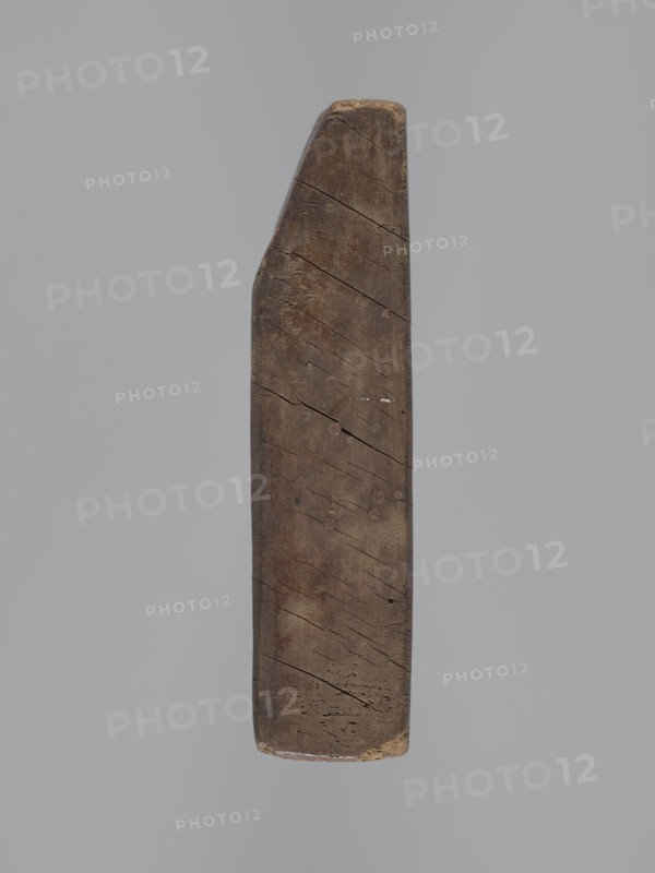 Double-sided gingerbread mould. Obverse: board with letters of the alphabet carved on it. Reverse: image of a running wild boar across a meadow. Rectangular double-sided mould; on one edge a date engraved: 1806, with the letter S between the digits on the background of an anchor mark of the woodcarver or gingerbread maker—owner of the form. Folk-style decoration, animals, wild boar iconography, alphabet. Material: wood form. Date: 1806. Place/style: Silesia Province of Silesia, 1742–1918. Dimensions: height 16 cm, width 12 cm, depth 3 cm., A single vertical wooden object with a weathered surface and a tapered top, showing a group of diagonal cracks and a few small circular marks, suspended against a plain light gray background; visible colors include brown tones on the wood and gray in the background.