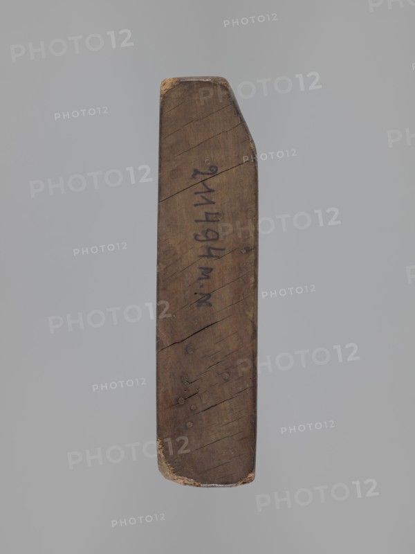 Double-sided gingerbread mould. Obverse: board with letters of the alphabet. Reverse: image of a running wild boar across a meadow. Double-sided rectangular mould; on one side a decorative board with the letters of the alphabet carved on it; the other side: image of a wild boar running across a meadow. On one of the edges there is a date engraved: 1806, between the digits of the date the letter S on the background of an anchor the mark of the woodcarver or gingerbread maker - the owner of the form. Materials/function: form for gingerbread; folk-style decorations; iconography includes wild boar and alphabet. Date: 1806. Place/region: Province of Silesia 1742–1918. Dimensions: height 16 cm, width 12 cm, depth 3 cm., A single elongated wooden object with a tapered end suspended against a neutral gray background, showing a weathered brown surface with visible wood grain, small cracks, and several round indentations; the top and bottom edges show worn areas with lighter beige exposed wood, ...