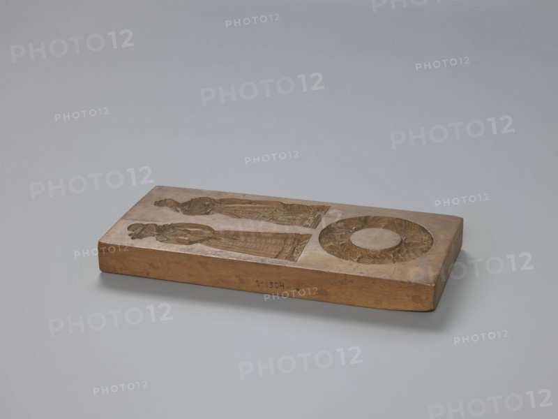 Double-sided gingerbread mould. Obverse: two female figures and a wreath; Reverse: two male figures and a house with a windmill. Woodcarving, folk-style decorations; genre scenes featuring houses, wreaths, windmills, peasants. Rectangular mould: one side—upper: two female figures in mid-19th-century costumes one in an elegant dress with a flower-decorated bonnet, the other in richly flowered folk dress; lower: a wreath woven of flowers and leaves. Other side—upper: two male figures in peasant costume left figure leaning on a shovel, right figure likely a sower; lower: a decorative motif of a house among meadows and trees with a windmill beside it. 18th–19th century, Province of Silesia 1742–1918. Form: double-sided mould. Height 28.5 cm; width 13 cm; depth 3.6 cm., A rectangular wooden block rests on a smooth light gray surface against a pale gray background, the block showing a warm brown color with slightly darker brown carved areas; the top face features carved reliefs including a p...