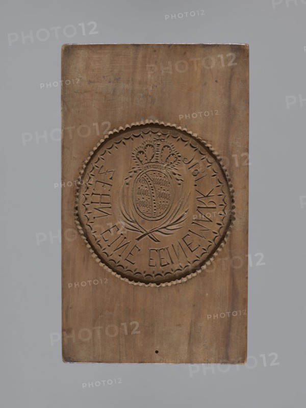 Double-sided gingerbread mould. Obverse: rectangular mould with an oval coat of arms in a decorative round frame topped with a crown; beneath the coat of arms two crossed palm leaves; around it an inscription in German: ZEHN EINE EEINE MARK 1816. Reverse: wreath woven from flowers and ears of grain. Gingerbread mould, folk-style decorative applied art, plant, geometric and floral ornaments. German, Silesia, ca. 1816. Form dimensions: height 15.7 cm, width 9 cm, depth 3.8 cm., A rectangular wooden block with a carved circular emblem in the center, showing a decorative scalloped border and an inner design featuring a crowned oval medallion surrounded by stylized leaves; the wood displays a natural brown color with variations in tone, a few darker brown marks and small spots, and the background around the block is gray.