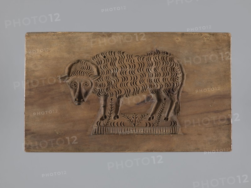 Double-sided gingerbread mold. Obverse: sheep; reverse: hare. Woodcarving, folk-style decorations; decorative and applied art; zoomorphic motifs, gingerbread molds. Rectangular mold: one side shows a sheep standing in a meadow with its head turned forward; the other shows a running hare with a flowery meadow at its feet. Iconography: hares, lamb. 18th–19th century, Province of Silesia 1742–1918. Mold. Height 15.5 cm; width 9 cm; depth 3.6 cm., A rectangular wooden panel with a carved relief of a sheep facing slightly left, showing a textured wavy fleece pattern, curled horns, a stylized face with carved eyes and snout, and slender legs standing on a decorative carved base; the wood surface shows grain variations and small knots, with visible tones of brown and tan on the panel and darker brown in the carved areas, set against a plain gray background.