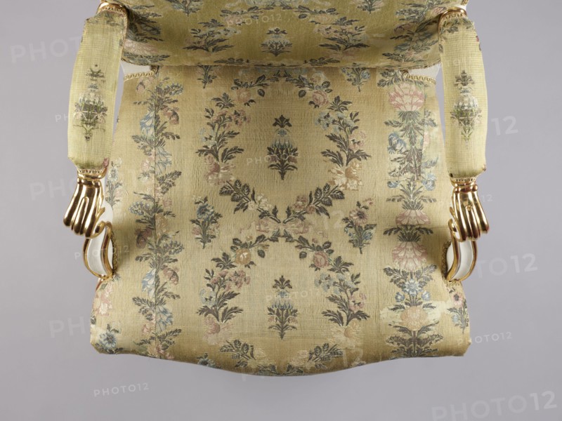 Armchair, Rococo style; floral decoration; cabriole legs; silk fabrics; 18th-century patterned floral fabrics. Upholstered seat, backrest, armrests; carved, white-lacquered and gilded. Cabriole legs arched into the frame, cut out in a wave-like manner at the bottom; trapezoidal seat with a slightly bulging front frame. Disc-shaped backrest, nearly square, topped with an arch, with slightly concave side rails, straight in line with the sitter's back and slightly tilted with a clearance. Armrests with a volute at the front end on recessed, slightly S-shaped supports. Edges of all frame elements highlighted with a gilded half-cylinder. Central part of the front frame and upper front legs with carved floral decorations; ends of armrests and feet with gilded acanthus leaves. Reupholstered with cream-colored silk fabric satin-weave ground, accented with colored silk and brocaded twill weave with silver thread; decorated with wreaths and alternating vertical garlands, with small floral sprigs...