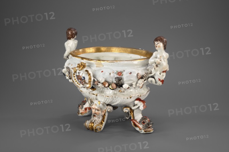 Vase from the Swan service, Koenigliche Porzellain Fabrique, Meissen 1737–1741. Makers: Johann Friedrich Eberlein 1695–1749, Johann Joachim Kaendler 1706–1775. Swan service; soup tureen form, oval vase on four modeled legs in the form of spits with plastic masks of bearded men or volutes—plastic dolphins with raised bodies and tails supporting the vase two. Two handles at the short ends in the form of fully modeled naked children—tritons with double decoratively crossed fish tails. Belly is knobbed and modeled with a shell motif in the lower part and a wave band in the upper part, with applied plastic flowers, polychrome-painted shells and seafood, and two oval shields in rocaille-moulure cartouches bearing a marital coat of arms supported by heraldic lions with jewels. Central underside features a convex gilded shell oval. No cover. Meissen, Saxony. Vessel; height 24.0 cm, width 34.0 cm., A decorative porcelain bowl sits against a gray background, primarily white with gold accents alo...