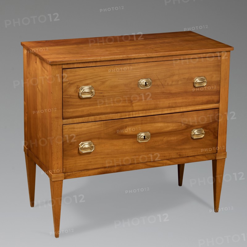Chest of drawers, Biedermeier style. Cherry, two-drawer cuboid chest on straight, tall, angular, tapering legs, polished. The body has a stand-alone structure with boarded sides and backs set in grooves; no bottom. Boarded drawer faces, recessed bottoms, with glued-on oak slides. Locks and handles of embossed brass sheet, rectangular with chamfered corners and an oval cabochon in the center; geometric cast brass pulls. Steel extruded single-bolt locks. 1820–1830, Southern Germany. Furniture storage, H 80.5 cm × W 87 cm × D 42.6 cm., A wooden chest of drawers with a warm honey-brown finish standing on tapered legs, featuring two large drawer fronts each fitted with several brass-colored oval handles and keyhole escutcheons, the wood grain visible across the top, sides, and drawer faces, and the piece set against a neutral gray background with soft shadows underneath; visible colors include brown, brass, gold, gray, and hints of darker brown in the wood grain.