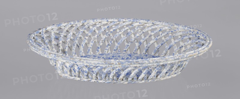 Openwork basket, Cmielów faience and porcelain factory founded 1804. Openwork basket with an oval, mandorla-like cross-section and parted walls formed by two layers of rollers rotating diagonally in opposite directions. Top enclosed in a band of three rollers; bottom finished in a braided-rope form. Interior bottom fitted with an oval plate. Surface decorated with a blue wallpaper pattern. Abrasions to the glaze and slight chipping on the rim. Dated 1840–1860. Poland; Cmielów. Ceramic vessel. Height 6.5 cm; length 30.5 cm; width 18.5 cm., A shallow oval woven basket with an open lattice structure made from twisted strips, covered in a patterned surface showing white and blue as the dominant colors with small areas of beige and faint gray; the weave creates curved diagonal openings around the sides and a flat base with a floral-like motif, all set against a plain light gray background.