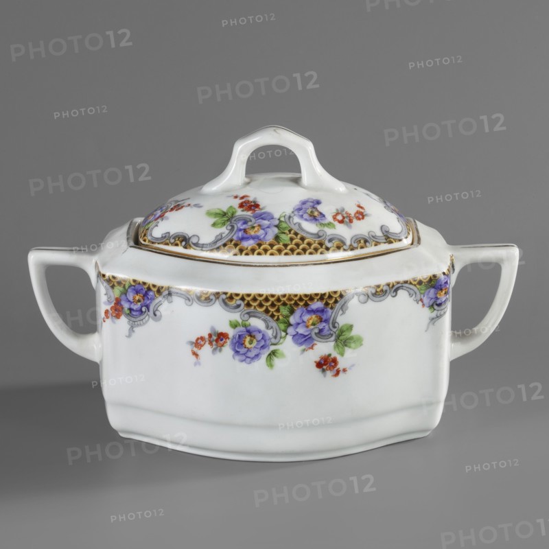 Sugar bowl with lid, Lwów model, Cmielów faience and porcelain factory est. 1804. Floral decorations; sugar bowl with wavy walls beveled on the sides, oval cross-section, with perpendicular handles beveled at the top. Slightly domed lid with a handle-shaped finial. Around the upper edge of the sugar bowl and lid is colorful tracing paper decoration with a fish-scale motif in rococo grey frames and sprigs of purple and brick-red flowers; around the rim of the vessel and the tops of the handles—gilded borders. Tracing paper technique. 1920–1930. Poland; Cmielów. Vessel. Height 11.5 cm, width 18.5 cm, depth 11.5 cm., A white porcelain lidded dish with a rounded rectangular body and a curved handle on each side, decorated with floral motifs in purple, red, green, and touches of yellow, a decorative band in gold with gray scrollwork along the upper edge, and a white loop handle on the lid; the background is gray.