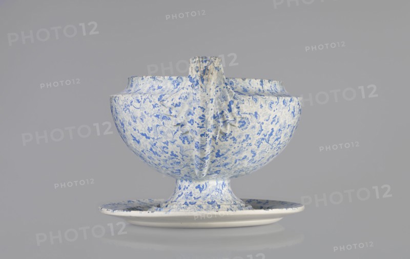 Vase, Cmielów faience and porcelain factory; 1804–, factory. Wallpaper pattern decoration, orchids, plant and floral decoration. Unknown provenance. A small vase with a convex belly supported by a small foot. Decoration in blue print with a wallpaper pattern. Vertical, fan-shaped handles, slender in a fantastic leaf shape with a line gently diverging from the belly and curling high towards the rim, surrounding the upper part of the vase with a fluted leaf. 1840–1860. Poland, Cmielów. Vessel. Height 16 cm; length 32 cm; width 20 cm., A ceramic bowl with a matching saucer, both covered in a dense floral pattern in blue on a white background; the bowl sits on a short pedestal foot and has a vertical handle-like ridge running down one side, with the blue floral motifs varying in intensity across the curved surfaces; the saucer mirrors the same blue-on-white pattern and sits beneath the bowl, and the overall scene is set against a smooth light gray background.