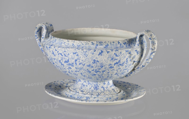 Vase, Cmielów faience and porcelain factory; 1804–, wallpaper patterns, orchids, plant decoration, floral decoration, unknown provenance. A small vase with a convex belly supported by a small foot. Decoration in blue print with a wallpaper pattern. Vertical, fan-shaped handles, slender in a fantastic leaf shape with a line gently diverging from the belly and curling high towards the rim, surrounding the upper part of the vase with a fluted leaf. 1840–1860, Poland, Cmielów. Vessel; height 16 cm, length 32 cm, width 20 cm., A large bowl-like vessel with two curved handles sits on a shallow matching base against a plain gray background; the surface of the vessel and base is covered in a dense floral and foliate pattern in shades of blue on a white ground, with the interior of the bowl appearing mostly white with a few blue specks, and the handles showing the same blue-and-white decoration.