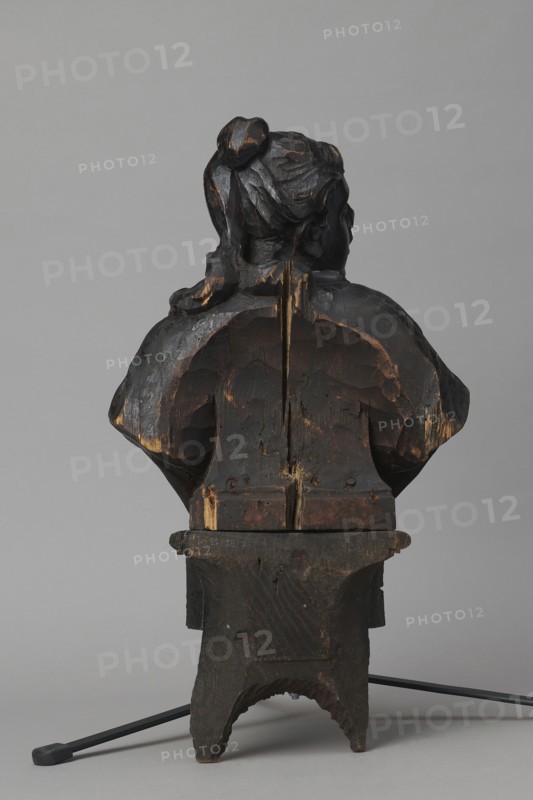 Bed finial from the Hospital of St. John of God in Warsaw, Fontana, Jakub 1710–1773 designer; co-creator, Redler, Jan Chryzostom 1704–1779 woodcarver; author; female busts; Hospital of St. John of God Warsaw; 1650–1944; Hospitallers of St. John of God order; Czartoryski, August Aleksander 1697–1782; baroque style. HOSPITAL BED FINIAL in the form of a pediment with volutes on the sides and a corbel in the middle supporting the bust of a young woman; fastened with two pins to the front section of the cornice crowning the canopy. The outline of the finial and the corbel are highlighted with a black ribbon stucco inlay. The corbel has an oval opening filled with a wooden, rotating medallion with inlaid letters on the obverse and reverse missing. The runners feature garlands made of ribbons and bay leaves; the volutes feature small brackets with turned, cup-shaped pinnacles. ca. 1757–1760, Warsaw; furniture fragment. Height 66 cm, width 93 cm, depth 16 cm., A carved wooden bust shown from t...
