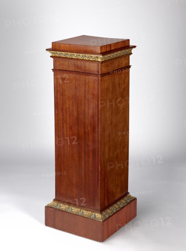 Pedestal cabinet, single-leaf with a drawer at the top, supported by a prominent plinth; veneered with mahogany, with profiled, carved, gilded and painted cornices and applied decoration on the doors. The edges of the door leaf and drawer faces and the corresponding front edges of the walls are diagonally cut. The molding around the plinth is decorated with a dark green and gilded leaf ornament; the cornice under the top is decorated with rosettes set in a rhomboidal grid. The upper section of the door features a bas-relief woman's head, below it a lotus flower surrounding the keyhole; the drawer face has a gilded wooden handle in the shape of a mushroom. Originally four shelves inside; the drawer bottom is recessed from the back. A single-bolt, self-locking steel mortise-and-apply lock and brass pintle hinges. Empire style; first quarter of the 19th century; possible origin Poland or Austria. Height 97.2 cm, width 33.5 cm, depth 35.2 cm., A tall wooden pedestal with a rectangular colu...