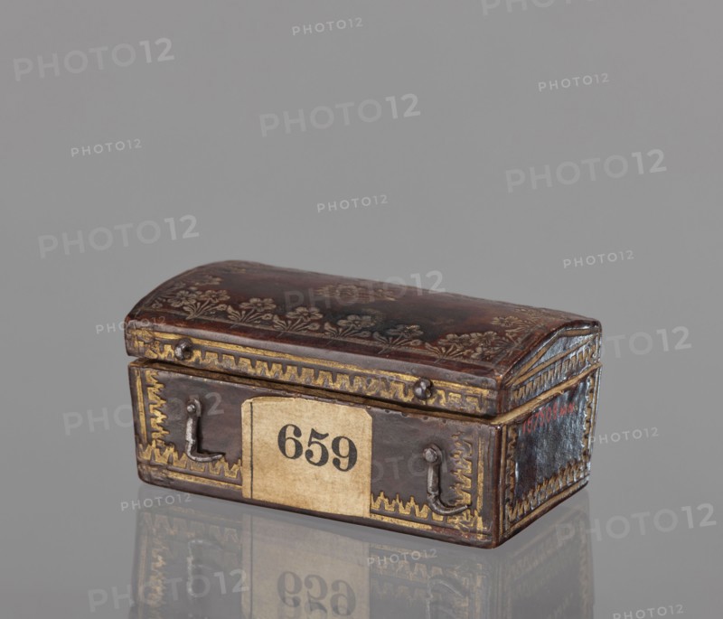 Case for a stamp, bookbinding, embossing; leather case in the form of a chest slightly widened at the top, with an eternal hinge slightly convex, covered with leather with gold-embossed decoration stripes and zigzag lines at the edges; on the lid, flowers with stems at the edge and one in the center. Interior lined with pink satin. 2nd half of the 18th century, Poland . Case; height 3.5 cm, width 7.8 cm., A small rectangular box with a domed lid sits on a reflective surface, showing a dark brown leather-like finish with decorative floral embossing on the top and gold-colored ornamental borders along the edges; the front and sides feature metal loop handles and corner embellishments, and the overall color palette includes dark brown, gold, beige, and hints of gray and reddish tones.