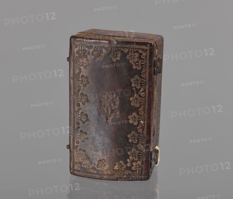 Case for a stamp, bookbinding, embossing; leather case shaped like a chest slightly widened at the top, with an eternal hinge slightly convex, covered with leather with gold-embossed decoration stripes and zigzag lines at the edges; on the lid, flowers with stems at the edge and one in the center. Interior lined with pink satin. 2nd half of the 18th century, Poland . Case; height 3.5 cm, width 7.8 cm., A rectangular box with rounded edges standing upright, covered in dark brown material with a worn, slightly glossy surface and a pattern of embossed floral motifs around the edges and a central flower design; the floral embossing shows lighter brown and muted gold highlights. The box has a visible hinge and small metal clasp on one side with a hint of dull metallic color, and the top edge shows lighter brown wear and a thin gold trim. The background is a uniform mid-gray, and the box sits on a reflective surface that creates a faint mirror of its lower edge.