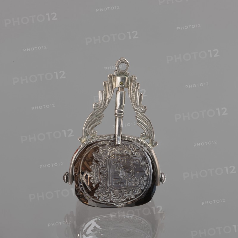 Seal stamp with the Pomian coat of arms; coats of arms; Pomian coat of arms; private seals; coat of arms seals; Axe coat of arms. Seal stamp made of smoke crystal, three-sided with three Polish coats of arms; silver binding with a volute motif. On individual fields: the Bawól head coat of arms; the Pomian coat of arms; a five-field coat of arms: inside Pomian, in the upper two Toxe, an unrecognized crowned rooster, in the lower ones: Rolbiecki and unrecognized. All under noble crowns, with jewels; the five-field surrounded by war trophies. 3rd quarter of the 18th century, Poland; stamp; height 5.2 cm, width 3.5 cm., A small decorative object with metallic silver ornamental fittings and a smoky brown faceted central stone, shown against a plain light gray background; the metal has a polished silver appearance with ornate scrollwork and a loop at the top, and the brown stone has a reflective surface and carved or engraved decorative patterns visible on its face, with a faint reflection o...