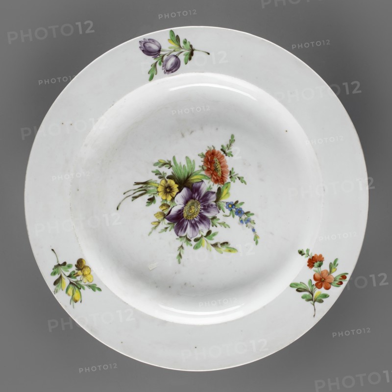Round platter, Korzec Porcelain and Faience Manufactory 1790–1832. Shallow plate with a wide, flat collar. White porcelain painted with floral motifs: collar has three sprigs with flowers in yellow, purple, and dark vermillion; the mirror shows a large bouquet with a purple anemone and a dark vermillion aster. Muted, cool palette — lemon yellow, dark purple, brown cinnabar — complemented by greens malachite and light yellow. Circa 1810–1815. Origin: Korzec, Volhynian Governorate, Poland. Vessel; height 5.7 cm, diameter 35 cm., A round white ceramic plate with a shallow well, shown against a gray background, decorated with a central floral spray featuring a large purple flower with a yellow center, surrounded by orange, yellow, and small blue blossoms and green leaves, and with additional small floral sprigs near the rim in purple and green, yellow and green, and orange and green; the plate shows subtle surface marks and light reflections.