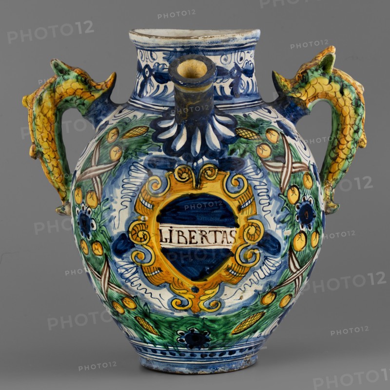 Pharmacy jug, Montelupo ceramics center; ca. 1400–, role classification of maker, pottery inscriptions — dragons, monograms, cartouches, plant decorations, jugs. A two-handled, ovoid pharmacy vase with a cylindrical neck, gently flaring at the rim. On the belly is a tubular spout, and on the sides are two handles in the form of fanciful dolphins dragons?. On the front is an oval cartouche with the inscription LIBERTAS framed by a massive laurel garland. The remaining background is grey and white, painted in two tones of cobalt, with a densely coiled twig with tripartite leaves and flowers a foglie. The decoration is dominated by cobalt blue in varying degrees of saturation, yellow, and green with accents of ochre and purple. Around the neck is a wavy twirl with birds. Below the handles is the letter B. The vase is an interesting example of Montelupo production, specializing in pharmacy vessels with leaf decoration inspired by Venetian ornamentation. Circa 1575, Italy, Montelupo, vessel...
