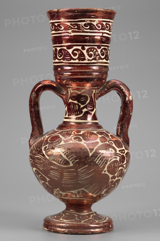 Amphora-type vase, Manises ceramic center; ca. 1400- author's role classification, pottery workshop author; mirrors decoration, birds, plant decorations. A slender, two-handled vase resembling a small amphora with a wide, long neck ending in a cup, and prominent, cylindrical, semicircular handles. The spherical body rests on a low spool foot. Painted with red mirror on a white background, with irregular patterns including a motif of exotic birds called pardalots and plant ornamentation. Horizontal trails with volute ornament on the neck. 17th century, Spain; Valencia; Manises. Vessel; height 26.5 cm, width 13.0 cm., An ornate vessel with a rounded body, a narrow neck flaring to a wide rim, and two curved handles rising from the shoulder to the neck, all decorated with carved or painted swirling and wave-like motifs; the surface shows a reflective, metallic sheen in deep reddish-brown and copper tones contrasted with lighter cream-colored areas where the decorative patterns appear, and ...