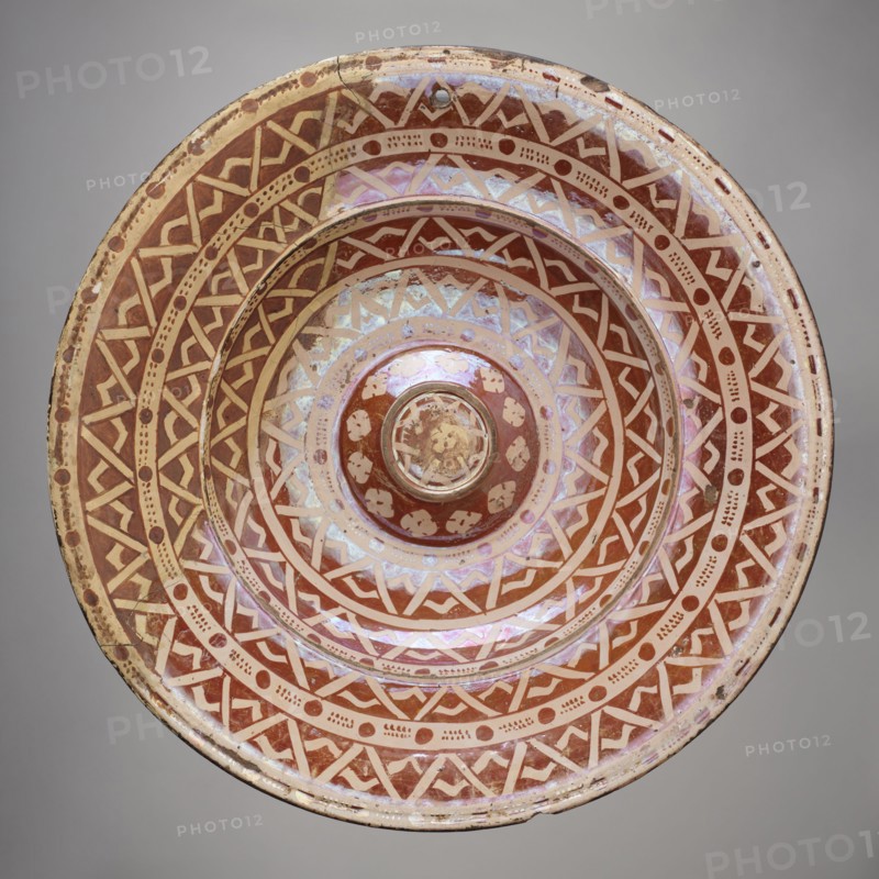 Platter, Manises ca. 1400–, Round ceramic platter with a wide collar and a bulging central cup-shaped bottom. Decorated with four concentric bands of varying widths filled with a geometric ornament resembling a braid composed of two intersecting zigzags; between them are white bands with an ornament of circles and dots. Around the central disc is a trail of four-leaf rosettes; a rosette in the center of the bottom. On the bottom: a wavy star, a spiral, and two rows of oval leaves at the edge. Geometric-floral ornament; 16th century, Spain Manises. Vessel; height 5 cm, width 40 cm., A round ceramic bowl with concentric decorative bands featuring geometric patterns and dot motifs in shades of reddish-brown and cream, showing an iridescent sheen with pale pink and purple highlights; the bowl has a raised central boss and visible wear with small chips and surface scratches; the background is a neutral light gray.