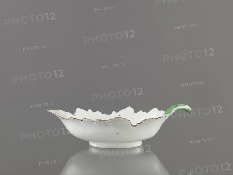 Leaf-shaped bowl, leaf-shaped bowl on a ring-shaped foot. Rims cut in the shape of leaves, triangular rim, on the opposite side a handle in the form of a green stem with veining. At the bottom is a bouquet with a large purple-pink flower, around it three small floral motifs. Four cracks extending from the rim, glaze abrasions. Porcelain, 1775–1800, Russia, Wierbilki; height 9.5 cm, width 12.0 cm, A shallow white ceramic dish with a scalloped rim showing worn brown edging and small speckles sits on a reflective surface against a gray background, with a curved green handle attached to one side.