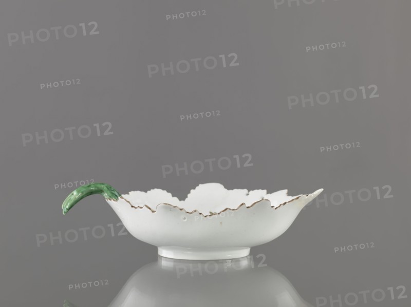 Leaf-shaped bowl, Wierbilki/near Moscow porcelain factory est. 1766. Maker: Gardner, Francis porcelain manufactory; 1766–1917. Leaf-shaped bowl on a ring-shaped foot. Rims cut in the shape of leaves with a triangular rim; opposite side a handle in the form of a green stem with veining. At the bottom a bouquet with a large purple-pink flower and three small floral motifs. Reconstructed tip of the spout; abrasions of the glaze. Date: 1775–1800. Place: Wierbilki, Russia. Vessel. Height 9.5 cm; width 12.0 cm., A shallow white ceramic dish with a scalloped rim showing a slightly irregular edge and a narrow rounded foot, sitting on a reflective gray surface against a smooth gray background; the rim has subtle brownish accents and one side of the dish features a curved handle glazed in green.