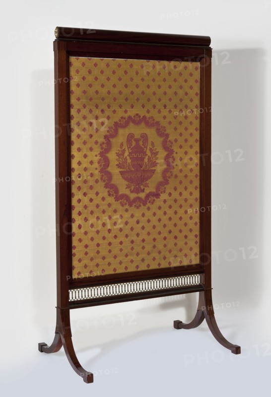 Fireplace screen, origin unknown, Empire style, wattle ornament, plant decoration, rosette ornament, decorative vases. Rectangular mahogany-veneered frame filled with patterned silk fabric, mounted on a pair of bipod supports. Posts and horizontal stiles are angular; the lower stile is an openwork brass strip with a wattle motif. Supports set in cubes are two arched, tapering angular legs ending in angular feet. Fabric stretched on an inner frame inserted from above into grooves of the side posts, enclosed by narrow slats; upper stile topped with a roller added during conservation replacing a non-original simy-profile strip. Brass rosettes on the sides of the shaft. Fabric: yellow plain-weave ground with a red satin pattern originally reversed color arrangement: central oval field with a vase on a pedestal framed by twigs; remaining field filled with small rosettes. Reverse upholstered with yellow silk fabric previously cherry-red linen canvas. A flat steel spring inside one side frame...