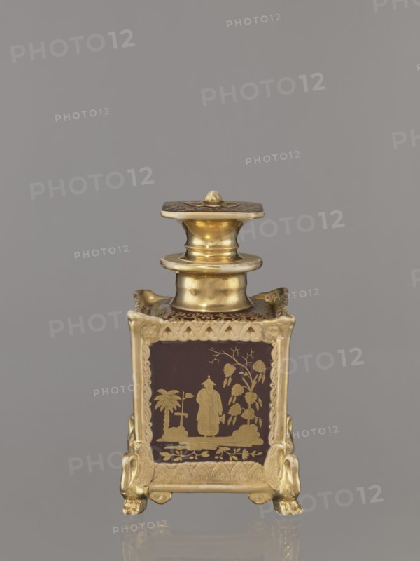 Bottle with lid. Porcelain manufactory of Francis Gardner 1766–1917. Chinoiserie cubic bottle with a profiled short neck and square stopper. Plastic ornamentation on the edges; swan-shaped feet in late Empire style one paw fragment missing. Gold-painted decoration with four genre scenes of Chinese people on the four sides; floral ornament on top of bottle and stopper; gilding abrasions. c. 1840, Russia; Wierbilki. Vessel. Height 18.5 cm; width 11.2 cm., A small ornate metal container stands against a plain gray background, featuring a polished gold-toned finish and a square body with a decorative lid and neck. The body sits on a base with four stylized feet shaped like animal paws, each foot and the frame around the panels showing intricate raised scrollwork in gold. Each visible panel has a dark brown background with a gold decorative scene depicting a standing figure in traditional attire beside stylized trees and foliage, framed by a gold border with repeating curved motifs. The lid...