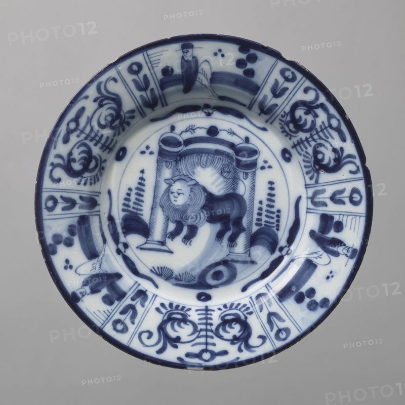 Plate, De Porceleyne Claeuw faience manufacturer; 1658–1840 manufacturer, animals, lions, plant decorations, chinoiserie. Semi-deep plate, quite thick-walled with a collar thinner at the edge. On a light blue background, decoration in dark cobalt. At the bottom, between two arbor posts flanked by schematically outlined bushes, a standing lion with a human face. The collar features six trapezoidal segments separated by narrow vertical fields with floral decoration, three of which depict a seated male figure, the other three arranged alternately with a schematic, symmetrical volute-floral composition. The lion at the bottom likely represents the Batavian Lion, symbolizing the House of Orange. 18th century, Netherlands, Delft; vessel; height 3.0 cm, width 23.2 cm., A round ceramic plate with a white base and blue painted decoration, showing a central scene of a stylized animal with a mane standing beneath an architectural element with columns, surrounded by curved brushstrokes and decorat...