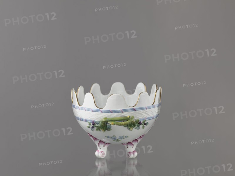 Vernier for glasses, Imperial Porcelain Factory in St. Petersburg 1744–1917. Oval vernier set on two volute-shaped arches with a serrated edge; below, a strip of relief basketwork framed top and bottom by a ribbon of three rods tied at regular intervals with a diagonal ribbon. The belly features painted colorful floral decoration and gilding. c. 1780, Russia, St. Petersburg. Vessel; height 16.5 cm, width 32.5 cm, depth 18.0 cm., A white porcelain bowl with a scalloped rim edged in gold sits on a reflective surface against a gray background; the bowl has a woven texture band near the top with a thin blue stripe, painted floral decorations in green and purple around its body, a raised green vine-like handle element, and three curled feet with pink and purple accents.