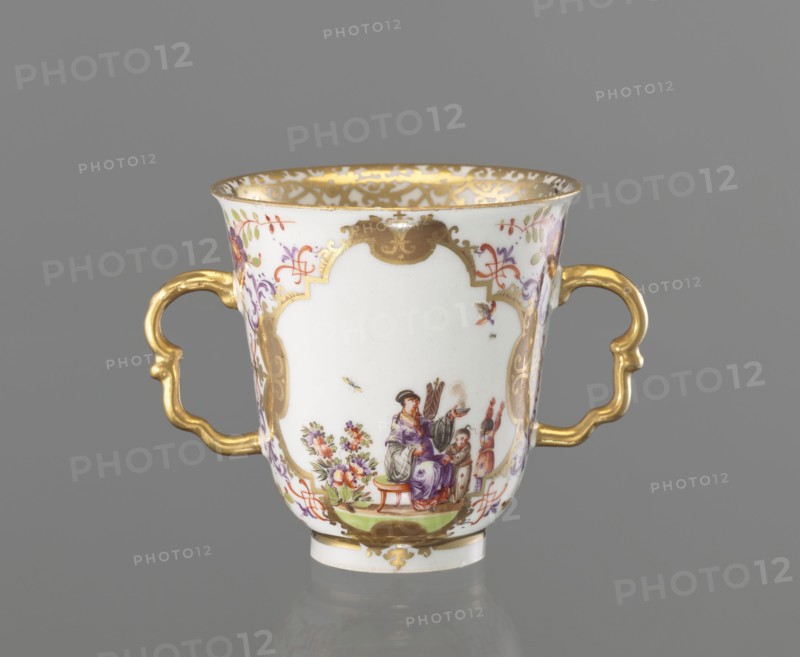 Two-handled cup, Koenigliche Porzellain Fabrique, Meissen ca. 1720–1735. Hoeroldtmalerei; chalice-shaped cup slightly parted at the rim with a rimmed foot and two two-part S-shaped gilded handles. Decoration: two four-part gilded cartouches on the bowl with a purple-brick-red pincer ornament; colorful miniatures of genre scenes with Chinese figures and plant motifs and insects: 1 a Chinese woman seated on a stool holding a fan and an incense burner with two Chinese children nearby; 2 a tea ceremony scene of a kneeling Chinese man with a cup and a standing man pouring water from a teapot, beside a table with utensils. Sides of the upper handles and beneath the foot painted with shoots of Indian flowers. Inner edge with a gold lace trail. Saxony, Meissen. Height 7.8 cm; diameter 7.5 cm., A white porcelain cup with two ornate gold handles and a gold rim sits against a gray background, decorated with gold cartouche shapes and delicate painted floral motifs in purple, red, pink, green, and ...