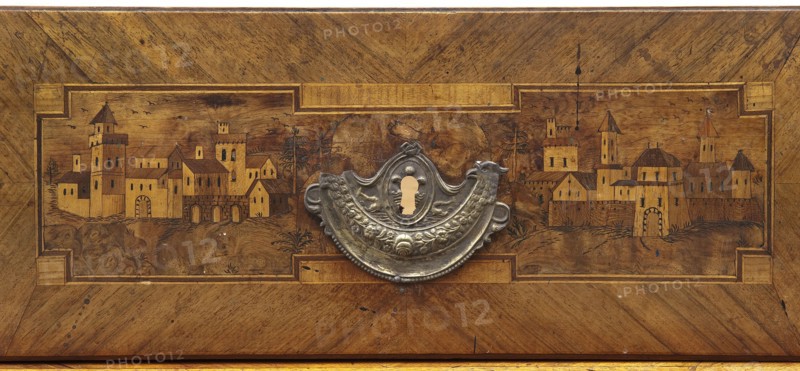 Chest of drawers; creator, revindication provenance; decorated with roses, vedute, dolphins, eagles, rosettes and geometric ornament in a classicism style; ca. 1790; Saxony; piece of furniture, storage piece; height 97 cm, width 143 cm, depth 62 cm, A wooden panel with a rectangular inset showing a stylized town scene carved or inlaid into the wood, featuring a group of buildings with towers, pitched roofs, arches, and small windows; a few trees and undulating ground surround the buildings and a few birds are sketched in the sky. Centered on the panel is an ornate metal escutcheon with a decorative, curved shape and embossed floral and scroll motifs, including a keyhole opening. The wood displays warm brown and golden tones with darker brown grain patterns and some small dark spots and blemishes, and the metal escutcheon appears silver-gray with areas of darker patina.