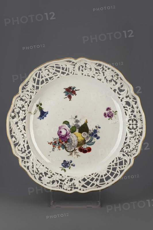 Platter with openwork, Königliche Porzellain-Fabrique, Meissen 1710–1763, ca. 1760. Round platter with a collar with a wavy gilded edge, cut into openwork with a motif of shell volutes and wavy flower branches, divided by two fan-shaped lattices with a flower or palmette motif; relief in the Bruehlsche Allerlei type. In the mirror, a colorful composition with a cut lemon, fig, cherry, currant and drop-shaped flowers, as well as projections of flower branches and a gooseberry branch. Porcelain, Meissen, Saxony. Vessel, height 6.7 cm, diameter 39.6 cm., A round white porcelain plate with a pierced ornate rim featuring floral and lattice cutwork and a thin gold edge, decorated in the center with a painted still life of fruit and flowers including purple, yellow, red, blue, green, and pink tones, with a few additional small floral sprays around the inner rim; the plate sits on a clear stand against a dark gray background.