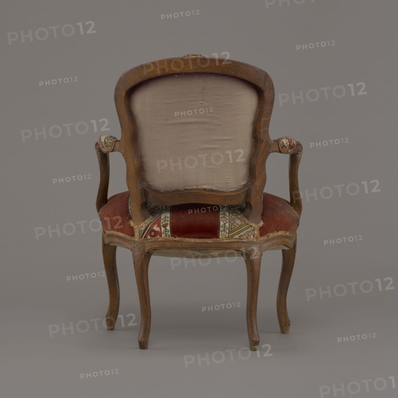 Armchair, rococo style, flowers and geometric ornament, ca. 1760, France; seating piece furniture, height 85.5 cm, width 62 cm, depth 60 cm., A wooden armchair shown from the back with a curved frame and carved top, a pale beige upholstered back panel, wooden curved arms with small padded rests covered in a patterned fabric, and a seat upholstered in worn red fabric with a decorative multi-colored patterned strip; the wooden legs are cabriole style with a natural brown finish, and the background and floor are uniform gray.