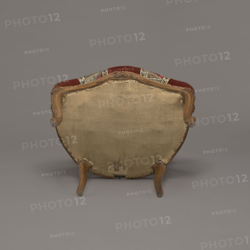 Armchair, rococo, flowers, geometric ornament, ca. 1760, France;, piece of furniture seating piece; height 85.5 cm, width 62 cm, depth 60 cm., An upholstered chair viewed from the back showing a beige fabric panel on the rear, a wooden frame with curved arm supports and legs in a brown wood tone, and a top edge with a strip of red upholstery and patterned fabric in colors including red, green, and cream; the background and floor are shades of gray.