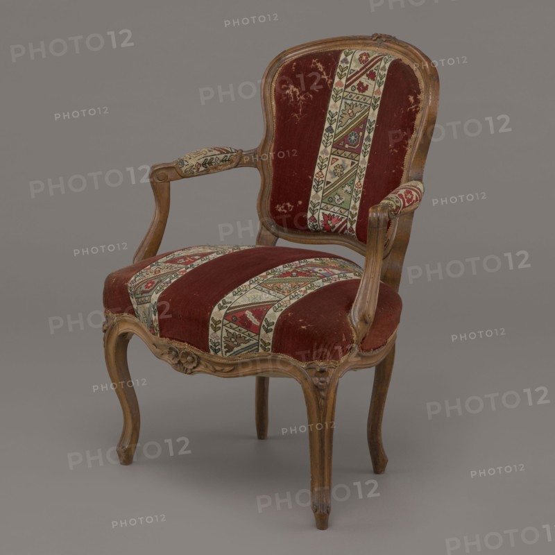 Armchair, rococo, flowers, geometric ornament, ca. 1760, France; piece of furniture seating piece; height 85.5 cm, width 62 cm, depth 60 cm., An upholstered wooden armchair with curved carved legs and a carved crest on the back, showing a worn deep red velvet fabric on the seat and back with patterned vertical bands of beige, green, red, and black tapestry; the wooden frame is a medium brown with visible grain and carved floral details, and the armrests have padded ends covered in the same patterned fabric; the background is a smooth neutral gray.