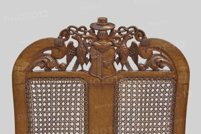 Armchair, provenance, Neo-Baroque, 2nd half of the 19th century, Western Europe. Baroque-style armchair with carved frame and woven rattan seat and back. Front cabriole legs terminate in claws clasping balls, decorated at the top with acanthus leaves; rear legs shaped like balusters terminating with ankles. Legs joined by an H stretcher with turned joints and side ankles. Trapezoidal seat with woven rattan and narrow frames with wavy cutouts at the bottom. High nearly rectangular backrest, S-shaped at the back, side rails curved inward at the top and topped with volutes. Center backrest divided by a narrow central board into two rattan-woven fields. Back crest features a two-field coat of arms supported by two eagles beneath a visor with labrets and a hat with wide brim and two cords with tassels. S-shaped armrests end in volutes at the front and rest on straight turned brackets set at an angle. Frame joints pegged. Furniture, seating piece; H 120 cm, W 70 cm, D 54 cm., A carved wooden...