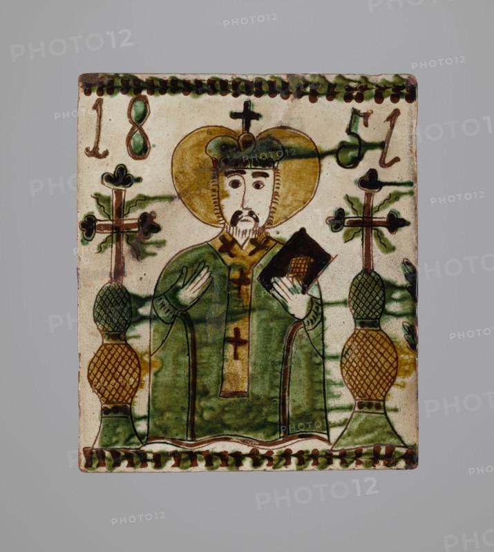 Tile, tiles, Hutsulshchyna, stoves, saints, date: 1851, with a representation of a saint, region: Hutsulshchyna, tile, height 23.7 cm, width 20.0 cm, depth 6.0 cm, A rectangular ceramic tile depicting a robed figure with a halo and a crown, holding a dark object in one hand and gesturing with the other, flanked by two tall decorative stands topped with cross-like elements; the figure's robe is predominantly green with brown and yellow accents, the halo is yellow-gold, and the crown is dark green with brown outlines, while the ceramic background is an off-white or cream color with green and brown decorative borders along the top and bottom and additional green streaks and brown outlines throughout.