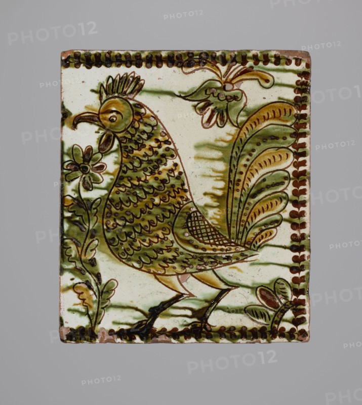 Tile, ceramic tiles with bird and plant decorations, stove tile with rooster motif, Hutsul region, 19th century, tile, height 20 cm, width 23 cm, depth 6 cm, A rectangular ceramic tile showing a stylized bird standing among decorative plants, rendered in shades of green, brown, yellow, and cream; the bird has a crested head, patterned body and layered tail feathers, with dark brown outlines and detailing, and there are a few floral and leafy motifs around it, a decorative border of alternating dark brown marks runs along the edges, and the background is mostly cream with green shading.