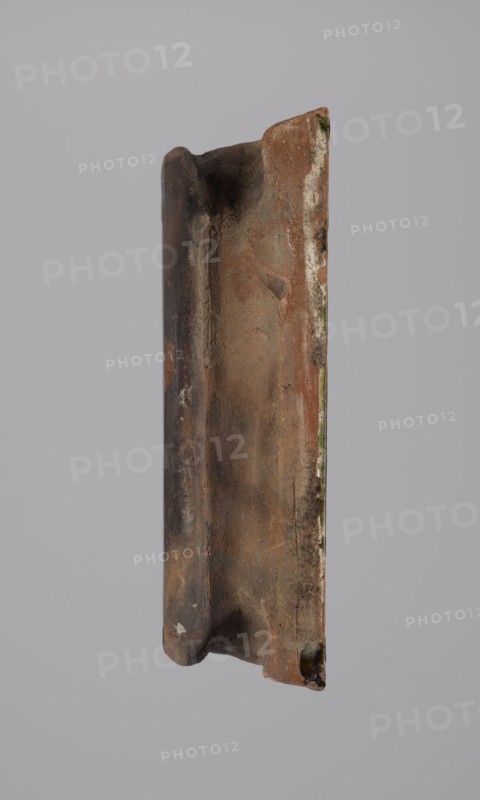 Tile, tiles, birds, stoves, plant decorations, Hutsul region, decoration with a rooster motif, 19th century, tile, height 20 cm, width 23 cm, depth 6 cm, A vertical weathered object with an uneven surface and rounded ridge, showing a range of earthy colors including brown, reddish-brown, dark brown, black, green, and small areas of pale off-white, set against a plain light gray background.
