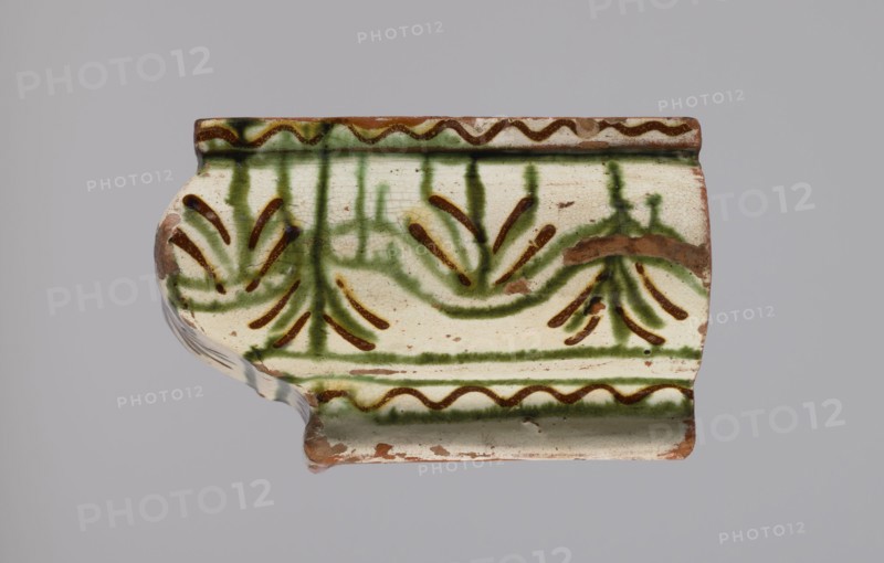 Cornice tile fragments 4, painted with a wavy twirl pattern; tile cullet; stoves; plant decorations; 19th century; Hutsul region, Poland., A fragment of a ceramic object rests against a plain light gray background, showing an off-white surface with green and brown painted decorative motifs; the piece has a curved profile with a raised rim along the top and bottom edges, the top rim featuring a wavy brown line bordered by green, and the body decorated with green vertical strokes and curved green bands accented by brown dash-like marks that form floral or fan-like shapes; there are areas of exposed reddish-brown clay where the glaze is chipped, and the glaze surface shows fine crackling and small dark specks.