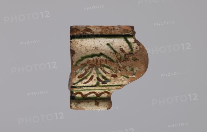 Cornice tile — fragments, four; maker, tile cullet; stoves; plant decorations; Hutsul region; tiles; painted with a wavy twirl pattern; 19th century; Hutsul region, Poland; tile cullet, A fragment of a ceramic object with a worn, irregular shape shows a surface of beige and light tan with areas of exposed reddish-brown clay; decorative brushstrokes in green and dark brown form curved and linear patterns across the face, and there are traces of darker green along raised ridges; the piece has a rounded protruding section on one side and a flat stepped edge on the opposite side, with patches of dirt and small chips visible on the surface against a pale gray background.