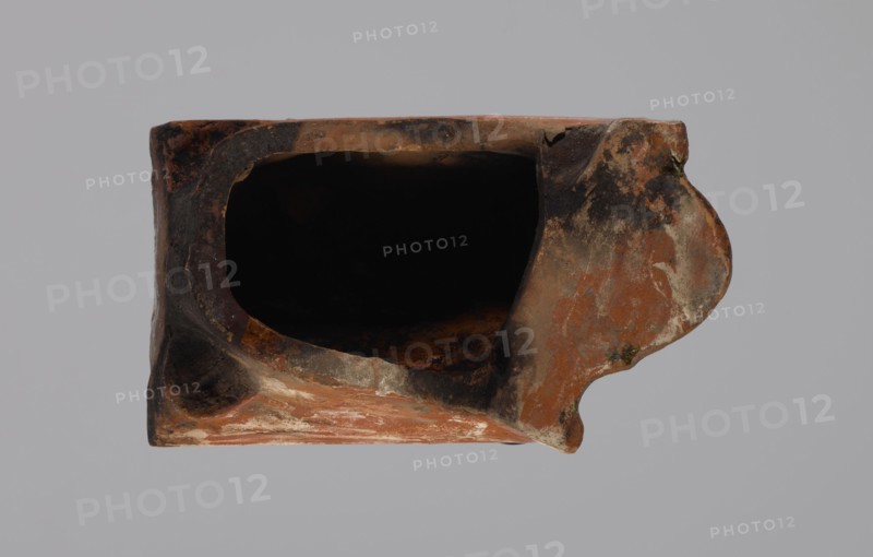 Cornice tile — fragments, four;, tile cullet; stoves; plant decorations; Hutsul region; tiles; Fragments of a cornice tile — painted with a wavy twirl pattern; 19th century; Hutsul region, Poland; tile cullet, A worn hollow object viewed from above with an irregular oval opening revealing a deep black interior, the exterior showing a textured surface with shades of reddish-brown, dark brown, black, gray, and patches of off-white, the form having a roughly rectangular body with an asymmetrical rounded protrusion on one side, edges and surfaces appearing chipped and rough with areas of discoloration and mottling.