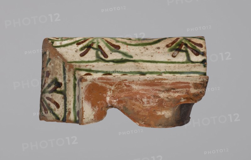 Cornice tile — fragments, four;, tile cullet; stoves; plant decorations; Hutsul region; fragments of a cornice tile painted with a wavy twirl pattern; 19th century; Poland., A broken ceramic fragment with a glazed surface showing white, green, brown, tan, and reddish-orange colors; the glaze includes green linear patterns and brown petal-like marks on a white background, with a worn reddish-orange terracotta area where the piece is fractured, and patches of tan and darker brown weathering visible across the surface.
