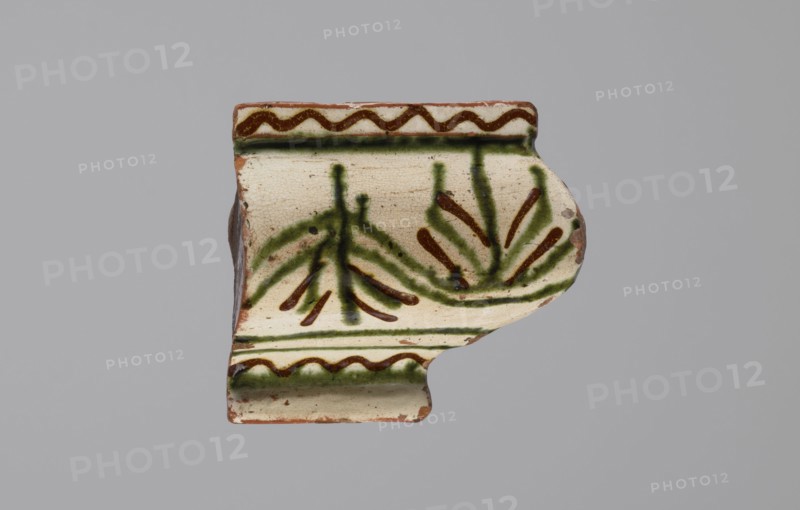Cornice tile fragments 4, tile cullet; painted with a wavy twirl pattern; 19th century; Hutsul region, Poland; stoves; plant decorations., A fragment of a ceramic tile with a cream-colored glazed surface showing a decorative pattern in green and brown; the shard has a curved edge and a molded profile with a raised band at the top and bottom, each band edged by a zigzag brown line and green shading, and the central field displays stylized plant-like motifs rendered in green strokes with brown accents; the ceramic body visible where glaze is chipped appears reddish-brown.