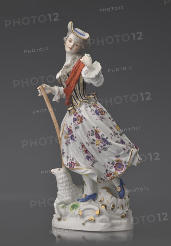 Shepherdess with a Sheep, Kaendler, Johann Joachim 1706–1775, sculptor; Koenigliche Porzellain Fabrique, Meissen 1710–1763, manufacturer. Figurine: On an oval base with rocaille and a tree trunk with a leafy branch — a young woman in a dancing running? pose, wearing a flowing flowered half-length skirt, a black-and-gold striped bodice with a small handbag tied at the back below a basque, a blouse with puffed sleeves tied at the elbow with a blue ribbon; a red sash with long ends thrown over the left shoulder and tied at the waist. Medium-length pinned-up hair falling in curls down her back, neckline, and shoulder; she wears a hat with a blue ribbon. She holds a shepherd’s staff upper part whipped resting on the head of a sheep lying at her feet; her head is tilted slightly left and she holds a flower whipped in her raised left hand. 19th/20th century after a model ca. 1750, Saxony; Meissen. Figurine, height 23.5 cm., A porcelain figurine of a woman wearing a small hat with blue and gol...