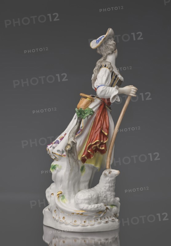 Shepherdess with a Sheep, Kaendler, Johann Joachim 1706–1775, sculptor; Koenigliche Porzellain Fabrique, Meissen 1710–1763, manufacturer. Porcelain figurine on an oval base with rocaille and a tree trunk with a leafy branch: a young woman in a dancing running? pose wearing a flowing flowered half-length skirt, black-and-gold striped bodice, small handbag tied at the back below a basque, blouse with puffed sleeves tied at the elbow with a blue ribbon; a red sash with long ends loosely thrown over the left shoulder and tied at the waist. Medium-length pinned-up hair falling in curls; she wears a hat with a blue ribbon. She holds a shepherd's staff upper part whipped resting on the head of a sheep lying at her feet. Her head is tilted slightly left and she holds a flower whipped in her raised left hand. 19th/20th century after a model ca. 1750, Saxony, Meissen. Figurine, height 23.5 cm., A porcelain figurine of a woman standing on a decorative base next to a reclining sheep, with the woma...