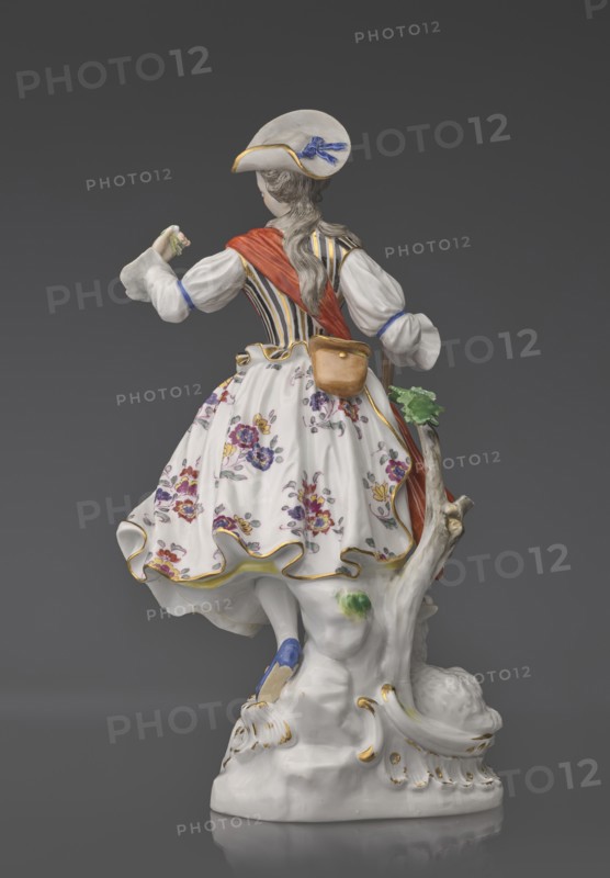 Shepherdess with a Sheep, Kaendler, Johann Joachim 1706–1775, sculptor; Koenigliche Porzellain Fabrique, Meissen 1710–1763, manufacturer. Figurine on an oval base with rocaille and a tree trunk with a leafy branch: a young woman in a dancing/running pose wearing a flowing flowered half-length skirt, a black-and-gold striped bodice with a small handbag tied at the back below a basque, a blouse with puffed sleeves tied at the elbow with a blue ribbon, and a red sash with long ends loosely thrown over the left shoulder and tied at the waist. Medium-length pinned-up hair falling in curls; she wears a hat with a blue ribbon. She holds a shepherd's staff upper part whipped resting on the head of a sheep lying at her feet; her head is tilted slightly left and she holds a flower whipped in her raised left hand. 19th/20th century after a model ca. 1750, Saxony; Meissen. Figurine, height 23.5 cm., A porcelain figure seen from behind wearing a wide-brimmed hat with a blue ribbon, a white blouse w...