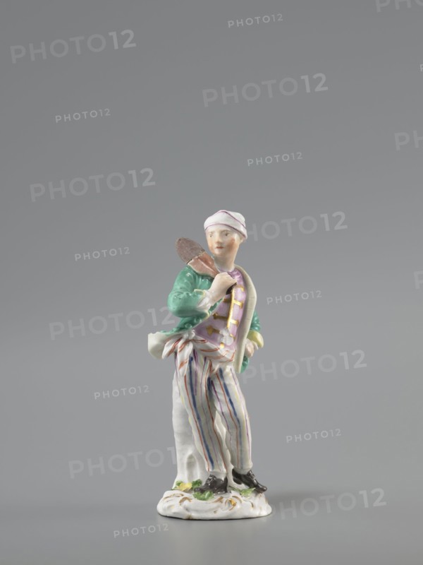 Man with a Potato Masher, Reinicke, Peter 1715–1768 modeler?, Koenigliche Porzellain Fabrique, Meissen 1710–1763 manufacturer, figurine. On a flat, circular pedestal with rocaille merging into a tree trunk, stands a young man leaning on the trunk at waist level. In contrapposto, head turned to the right, he holds a short oval board with a handle—an instrument for stewing potatoes—resting on his right shoulder. Dressed in trousers with colorful thin stripes tied loosely at the waist with a belt, loose ends, black shoes with a buckle, a short green jacket with box pleats, a pink waistcoat, with gold braiding and a soft cap similar to a nightcap. Ca. 1755, Saxony; Meissen; figurine; height 13.5 cm., A small painted porcelain figurine of a standing person wearing a white cap with pink trim, a green jacket, a pink vest, and white trousers with vertical blue and pink stripes, holding a brown object over one shoulder and leaning against a white tree stump, mounted on a white decorative base w...